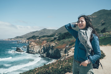 vintage style portrait of hipster young asian girl with backpack enjoying sunshine on peak mountain. female hiker looking sunlight flare in trip Big Sur rocky coastline USA california spring holiday