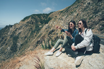 Two asian women friends taking break while trekking in mountain in big sur california usa. young girls hikers holding smart phone showing sharing pacific ocean partner relaxing on cliff near highway © PR Image Factory