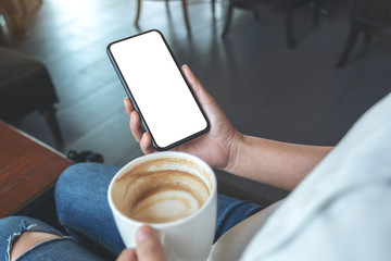 Mockup image of hands holding black mobile phone with blank screen while drinking coffee in modern cafe