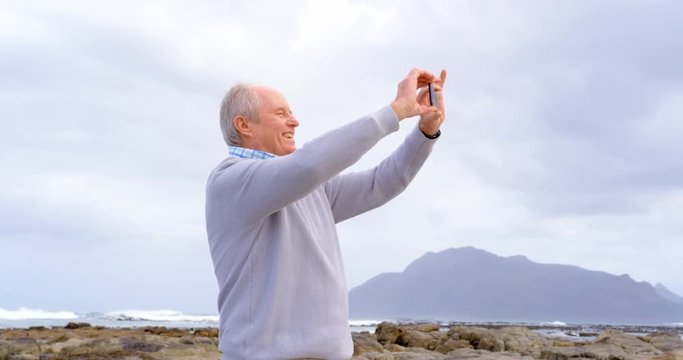 Front view of old caucasian senior man clicking photo with mobile phone at beach 4k