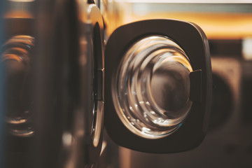 A dark yellowish room of a publiс laundry with a row of washing machines, selective focus on the opened door of one of the tumble dryers, shallow depth of field