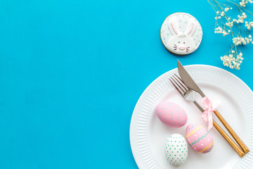Fototapeta na wymiar Easter dining table in pastel colors. Plate, cutlery, painted eggs, gingerbread and dry flower branch on blue background top view copy space