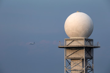 A weather radar tower with blue sky background and blured airplane