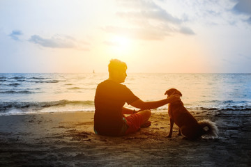 Man and dog sitting on sea beach and looking at sunset. Friendship and dedication between human and...