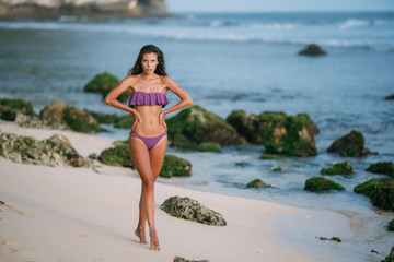 Sexy woman in purple colour swimsuit posing on sandy beach with big stones on background