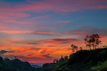 Dusk cloudy sky in evening. Red colorful sunset on top of hills with trees silhouette. 