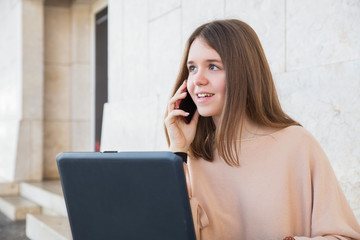 Positive female teenager using laptop and phone at building wall