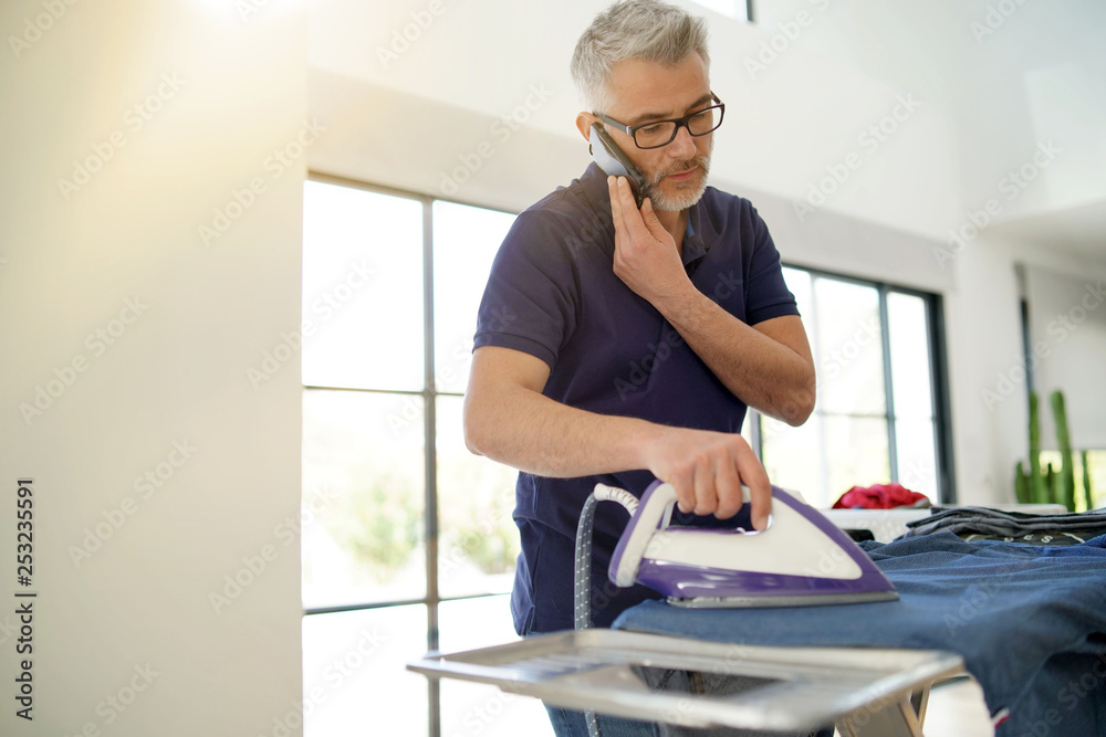 Poster mature man talking on cellphone whilst ironing shirt at home