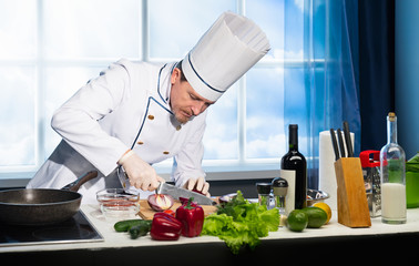 Cook in white uniform cuts red onion with knife on wooden board in the kitchen