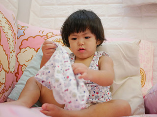 Fototapeta premium Little Asian baby girl, 23 months old, learning to put on short pants by herself - child development by allowing them to do things by themselves