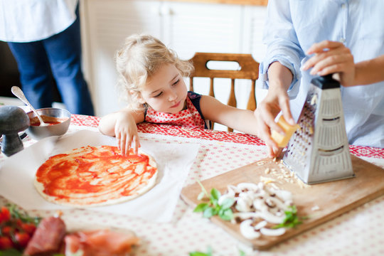 Family Is Cooking Homemade Pizza. Cute Kid Is Helping Mother To Grate Cheese. Funny Curious Child Is Interested In Preparing Italian Food And Meal In Cozy Kitchen. Lifestyle, Authentic, Candid Moment