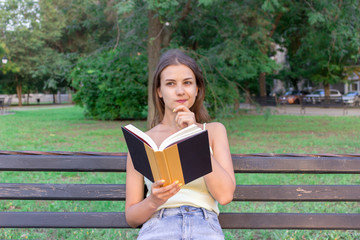 Beautiful woman is reading a book and thinking about something on the bench in the park. Girl has some thoughts and ideas