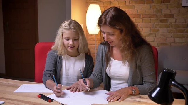 Closeup portrait of young mother and her little pretty daughter playing a tic tac toe game on the paper together at cozy home indoors