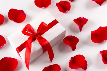 White gift box with red ribbon red velvet rose petals on white background. Flat lay