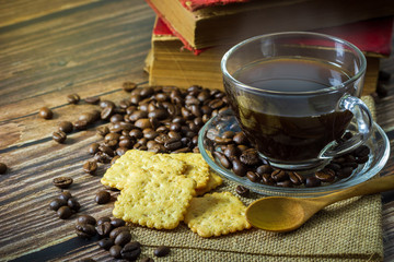 Naklejka premium Black coffee in clear glass cup and coffee beans with crackers and old book on wooden table.