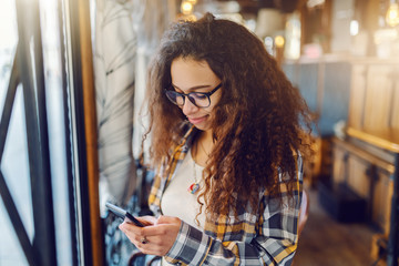Cute mixed race girl with curly hair standing in cafeteria and using smart phone.