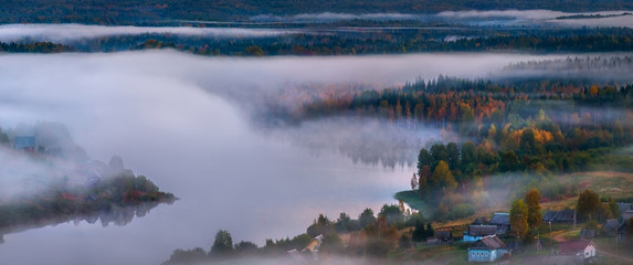 Foggy panorama of the village on the lake. Autumn dawn. Fog over the village, houses, village. On the shore of the lake in the fog.