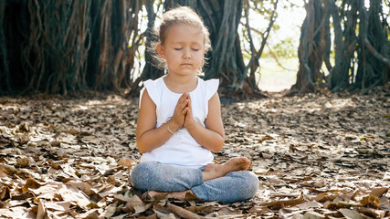 Little cute child girl meditating alone with crossed feet under banyan tree 