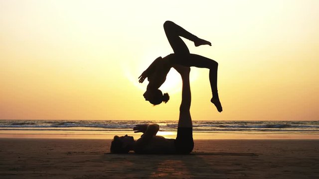Silhouette of fit sporty couple practicing acrobatic yoga with partner together on the beach in slow motion. Female acrobat balancing on feet of her partner in fylfot shape