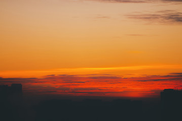 Cityscape with vivid fiery dawn. Amazing warm dramatic cloudy sky above dark silhouettes of city building roofs. Orange red sunlight. Atmospheric background of sunrise in overcast weather. Copy space.