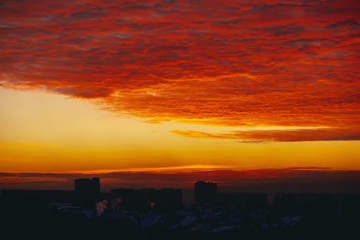 Cityscape with fiery blood vampire dawn. Amazing warm dramatic cloudy sky above dark silhouettes of city buildings. Orange sunlight. Atmospheric background of sunrise in overcast weather. Copy space.