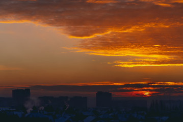 Cityscape with vivid fiery dawn. Amazing warm dramatic cloudy sky above dark silhouettes of city building roofs. Orange sunlight. Atmospheric background of sunrise in overcast weather. Copy space.