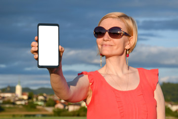 Beautiful woman wearing sun glasses while holding in hand a smartphone with a white screen on a countryside on a hot sunny summer day