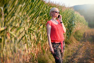 Naklejka na ściany i meble Young beautiful woman in colorful cloths and sunglasses outdoor talking on the cell phone with the corn field in the background and with one arm in the pocket