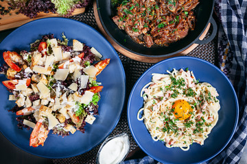 Top view on served food table. Caesar salad with shrimps, meat pan, carbonara on blue plate. Restaurant menu, recipe photo with copy space. Delicious flat lay food. European cuisine. Background image