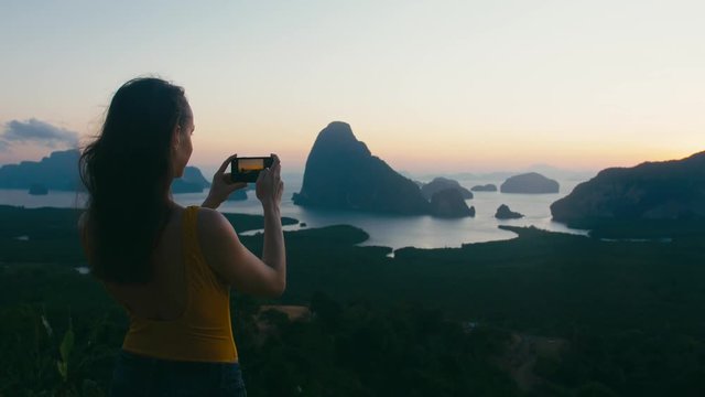 Woman tourist makes photo using smartphone at famous travel landmark viewpont Phang Nga bay in sunrise. Asia, Thailand