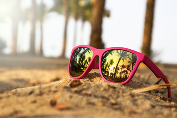 Pink modern sunglasses on the beach sand with palm trees and sunset reflection 