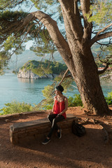 the girl sits on a trap under an old tree on a cliff with a beautiful view of the sea and mountains. Vacation in the European resort. Travel and leisure.