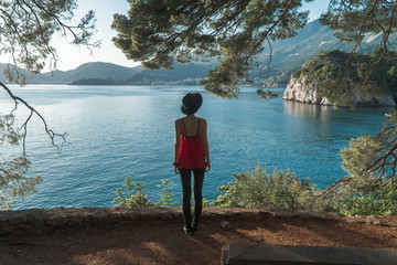 A girl stands under an old tree on a cliff with a beautiful view of the sea and mountains. Vacation in the European resort. Travel and leisure.