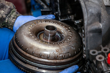 Close-up of a young male repairman in a working uniform repairing a torque converter and installing it in an automatic transmission of a supported car in an auto repair shop.  - obrazy, fototapety, plakaty