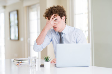 Naklejka premium Young business man working with computer laptop at the office peeking in shock covering face and eyes with hand, looking through fingers with embarrassed expression.