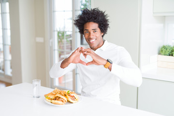 Fototapeta na wymiar African American hungry man eating hamburger for lunch smiling in love showing heart symbol and shape with hands. Romantic concept.
