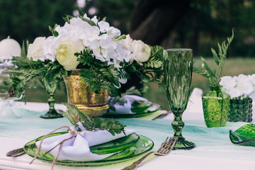 Wedding table setting with plates and green glasses decorated with white candles, green leaves and tablecloth outdoors