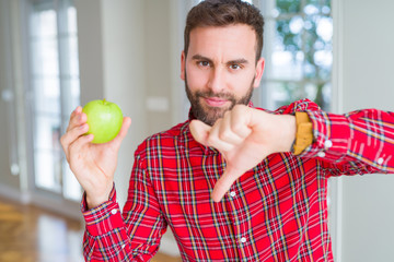 Handsome man eating fresh healthy green apple with angry face, negative sign showing dislike with thumbs down, rejection concept