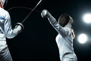 Young fencer athlete wearing mask and white fencing costume. holding the sword on black background with lights.