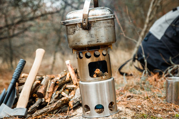 camping woodstove and utensils, axe and sapper shovel near a firewoods, backpack on the background