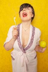 Plump woman with short black hair stands in a pink Bathrobe with teeth brush and green apple on a yellow Studio background alone