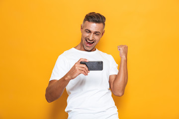 Emotional man posing isolated over yellow wall background play games mobile phone.