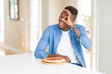 Fototapeta na wymiar African american man eating pepperoni pizza at home with happy face smiling doing ok sign with hand on eye looking through fingers