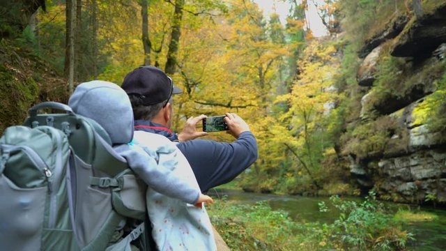 Hiker with little son in touristic backpack taking a photo of beautiful autumn forest 