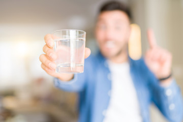 Young handsome man drinking a glass of water at home surprised with an idea or question pointing finger with happy face, number one