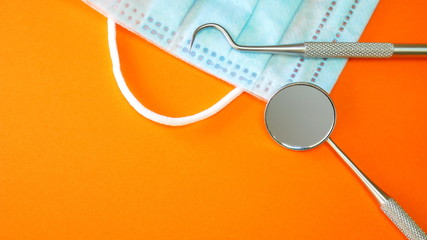 Dentist tools or instruments in dental office: dental explorer, dental mirror and surgical or procedure face mask on orange surface for copyspace text. Dental hygiene and health conceptual image