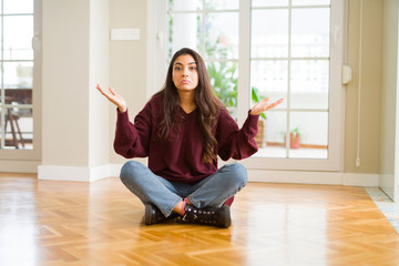 Young beautiful woman sitting on the floor at home clueless and confused expression with arms and...