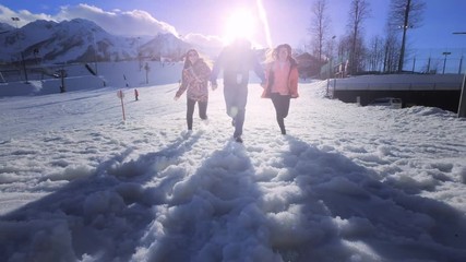 Company of friends having fun on holiday in the winter mountains. One guy and two girls enjoy the winter at the ski resort. They run in the snow, jump. Snow flies into the camera. Behind the mountain