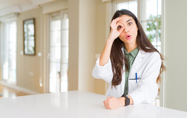 Fototapeta premium Young woman wearing medical coat at the clinic as therapist or doctor doing ok gesture shocked with surprised face, eye looking through fingers. Unbelieving expression.