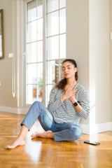 Fototapeta na wymiar Young beautiful woman sitting on the floor at home smiling with hands on chest with closed eyes and grateful gesture on face. Health concept.